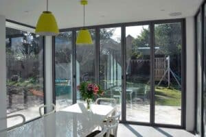 Sunlit dining area featuring glass walls beneath a flat roof extension with an overhang, showcasing a white table, yellow pendant lights, and a vase of flowers. Outside, a garden with trees, lawn, and play structure is visible.