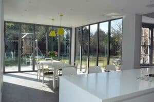 Modern kitchen and dining area with large glass windows set in a flat roof extension with overhang, featuring white furniture and yellow light fixtures, overlooking a garden with a playground.