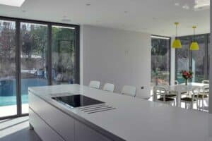 Modern kitchen and dining area with large windows under a flat roof extension with overhang, white counters, yellow pendant lights, and a view of the backyard pool.
