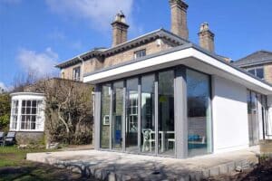 A modern flat roof extension with overhang is seamlessly attached to a brick house with chimneys, boasting large sliding doors that reveal a visible interior dining area.