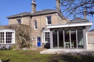 A large brick house with a modern flat roof extension, complete with an overhang and sleek glass design, featuring a blue door and surrounded by a lush garden.