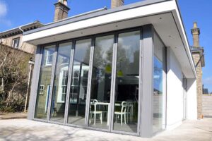 A modern flat roof extension with overhang showcases large glass doors and windows, featuring a cozy sitting area with white chairs and tables inside.