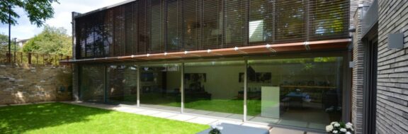 Modern house exterior with large glass windows and a sleek glass sliding door, complemented by wooden slat shading and a well-kept lawn in the foreground.