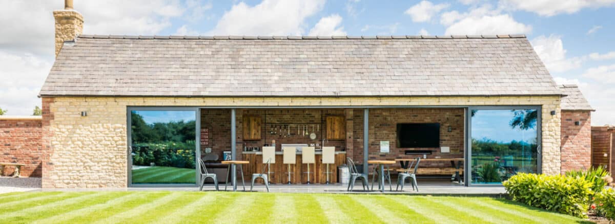 A modern outdoor bar and seating area in a stone building features sleek sliding aluminum doors, seamlessly connecting to the neatly mowed lawn and garden.