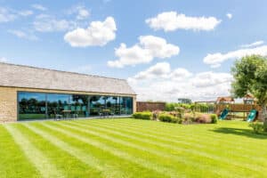 A modern building with large glass windows and elegant sliding aluminium doors on a sunny day, surrounded by a neatly mowed lawn, shrubs, and a playground area with slides.