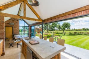 A bright, spacious kitchen features a concrete island and wooden beams, with large sliding aluminium doors that open to a lush garden and play area.