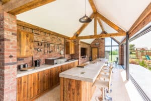 A spacious kitchen with a brick and wood interior features a concrete island with white stools, high wooden beams, and large sliding aluminium doors that open to a garden.