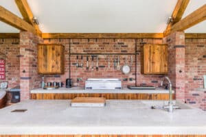 Outdoor kitchen with a brick wall, wooden cabinets, and countertop. Featuring sliding aluminium doors that open to reveal a grill, sink, and hanging utensils.