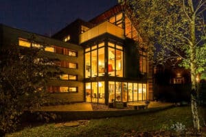 Modern house with aluminium bi-fold doors and large windows glowing warmly at night, surrounded by trees and a grassy lawn.