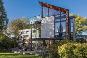 Modern three-story house with large glass windows and aluminium bi-fold doors, a slanted roof, and a patio featuring outdoor furniture, surrounded by greenery on a sunny day.