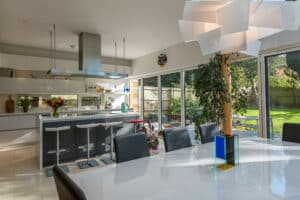 A modern kitchen and dining area features a large table, bar stools, a unique ceiling light, and large aluminium bi-fold doors that open to reveal the garden view.