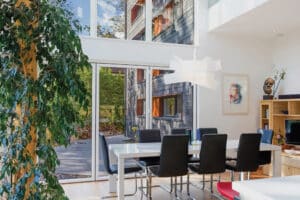 A modern dining room featuring a glazed gable end with a large window, a sleek white table, black chairs, a potted plant, and framed artwork on the wall.