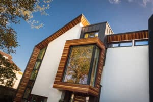 Modern house facade featuring aluminium bi-fold doors and a large wooden-framed window, with white walls and angled rooflines. It's set against a blue sky with some trees visible.