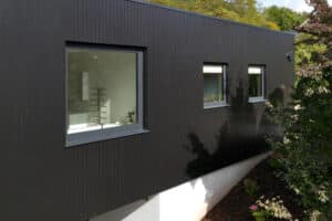 Modern black wooden building exterior with sleek corner windows and three rectangular ones, all complemented by lush surrounding greenery.