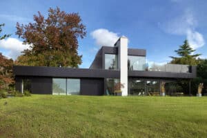 Modern two-story house with large glass and corner windows, surrounded by a grassy lawn and trees, under a partly cloudy sky.