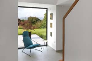 A modern, minimalist room features a blue chair and small table by corner windows overlooking a garden and hills. Three small paintings adorn the wall.