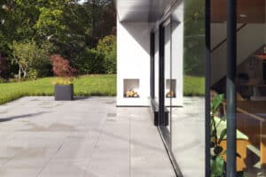 A modern house with large glass and corner windows opens to a spacious patio. A neatly stacked woodpile is visible in a recessed area on the patio, and the background features a lush green lawn and trees.