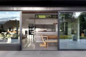 Modern kitchen and dining area with large glass doors, corner windows, gray countertops, wooden bench seating, and lush greenery outside.