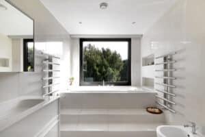 A modern bathroom with a bathtub next to corner windows overlooking green trees. It features white tiles, a sink, and a bidet, with chrome towel racks on the walls.
