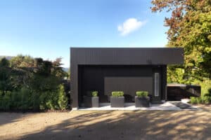 Modern black house with a flat roof, adorned with corner windows that invite natural light, sits elegantly amidst trees and greenery, featuring three potted plants at the entrance.