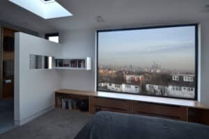A modern bedroom with a large window and 3 track sliding patio doors offering a stunning city skyline view. It features built-in shelves and a cozy bench beneath the window, while a skylight above floods the space with natural light.