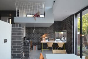 A modern interior with dark brick walls features a loft area with a net and someone lowering a rope. Below, 3 track sliding patio doors open to reveal a kitchen and workspace with two chairs, a table, and large windows.