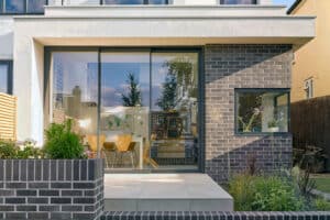 A modern house exterior showcases gray brickwork and a charming small garden. The highlight is the large 3-track sliding patio doors, offering a glimpse inside to a dining table and chairs.