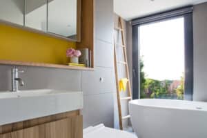 Modern bathroom with a freestanding tub, wooden accents, a yellow wall, and a tall window with 3 track sliding patio doors that open to lush greenery. A ladder shelf leans against the wall.