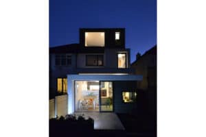 A modern, multi-story house at night features large windows and 3 track sliding patio doors illuminated from inside, showcasing a well-lit interior and a minimalist exterior design.