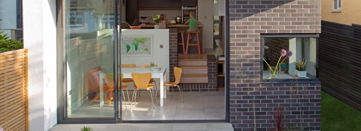 A modern dining area with yellow chairs and a table is visible through expansive 3 track sliding patio doors. A small window with a vase adorns the right brick wall, completing the stylish look.