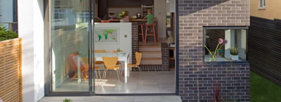 A modern dining area with yellow chairs and a table is visible through expansive 3 track sliding patio doors. A small window with a vase adorns the right brick wall, completing the stylish look.