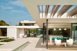 Modern house with large glass windows and sleek sliding doors, outdoor pool, and patio with seating area. White and wood elements dominate the architecture, with a minimalist design.