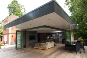 Modern house with a large open-concept living area, featuring corner bi-fold doors, a sofa, chairs, and kitchen. Outdoor deck surrounds the space, with trees in the background.