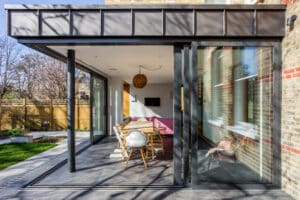 Modern glass-walled extension with corner bi-fold doors offers a view of a dining area featuring a wooden table, chairs, and a pendant light. The space seamlessly opens to a garden with a wooden fence.