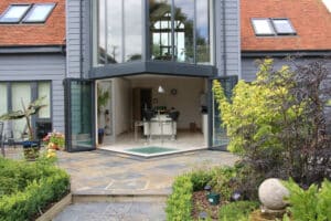 Modern house exterior with corner bi-fold glass doors open to a stylish kitchen. The patio features various plants and a stone pathway leading to the entrance.