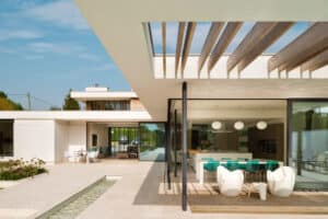 Modern house with open layout, large glass windows, and wooden beams. Features corner bi fold doors leading to outdoor seating and dining area, complete with a small water feature that sparkles on a sunny day.