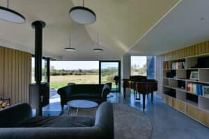 Modern living room with large corner bi-fold doors, two dark sofas, a round coffee table, a wood-burning stove, a grand piano, and built-in bookshelves. Experience the seamless transition between indoor comfort and the grassy landscape outside.