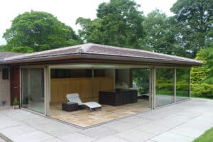 Modern patio with floor-to-ceiling glass walls and corner bi-fold doors, featuring a wicker seating arrangement and a lounge chair, surrounded by lush greenery.