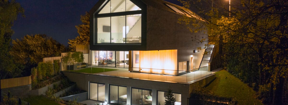 Modern two-story house with large windows and sliding aluminium doors, featuring an outdoor terrace illuminated at night, surrounded by trees and greenery.