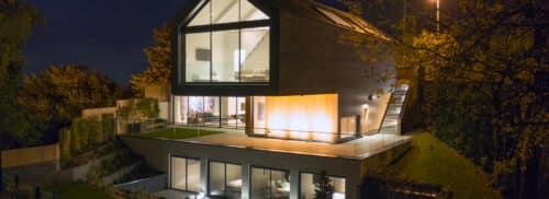 Modern two-story house with large windows and sliding aluminium doors, featuring an outdoor terrace illuminated at night, surrounded by trees and greenery.
