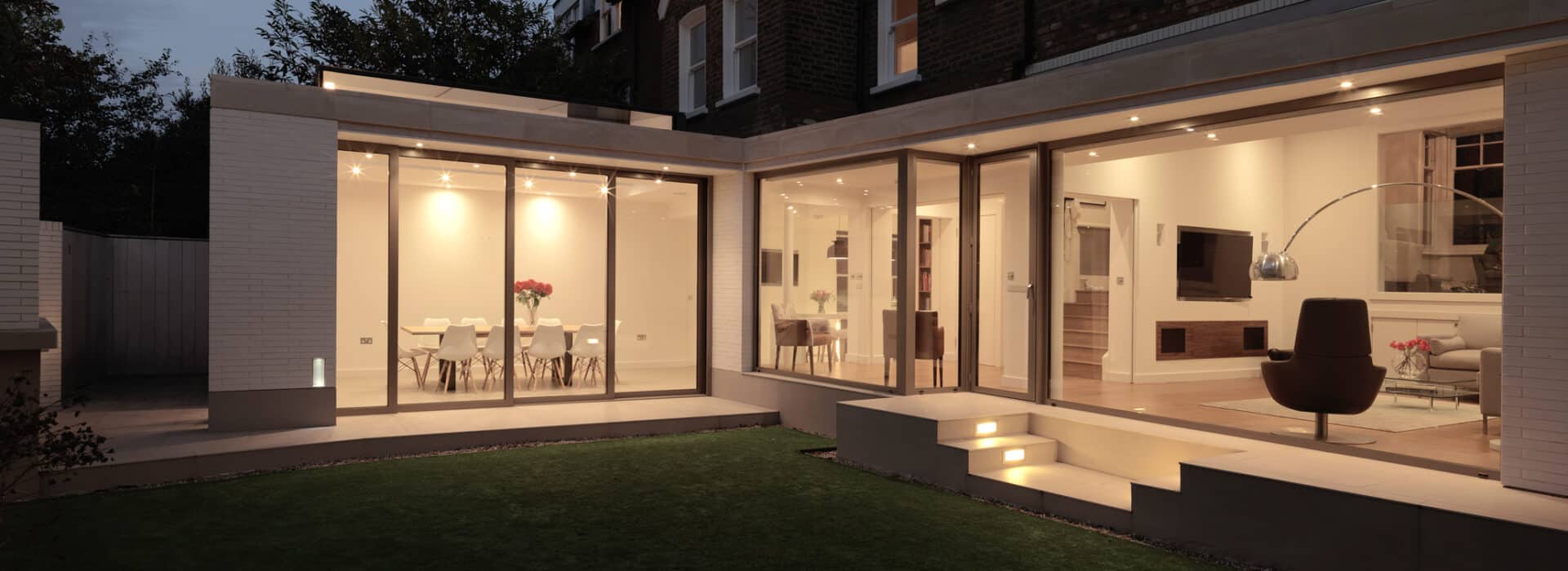 Modern house with large glass windows and sliding aluminium doors opening to a lit interior, showing a dining area with flowers and a living room, viewed from the backyard at dusk.