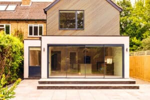 Modern house extension with large glass sliding doors, a brick exterior, and a sloped roof. The patio features gray tiles in the foreground, complemented by lush greenery around the sides.