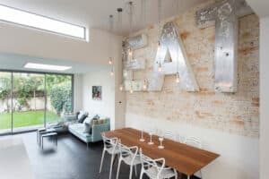 Modern dining area with large metallic "EAT" sign on a brick wall. Features a wooden table and white chairs. Adjacent living space with a couch and glass sliding doors overlooking a garden.