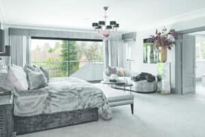 Luxurious bedroom with a plush bed, large windows, gray carpeting, and a chandelier. A glass sliding door opens to elegance, while sofa seating invites relaxation. Floral arrangement on a tall vase.