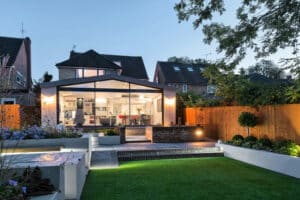 Modern house with glass extension and a sleek glass sliding door, landscaped garden, and patio at dusk. Indoor space is visible through large windows, surrounded by trees and fencing.