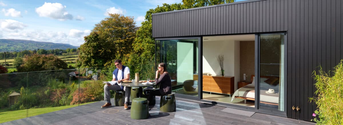 Two people sit at a table on a modern outdoor deck by a black house with large glass doors and striking corner windows, overlooking a scenic green landscape.