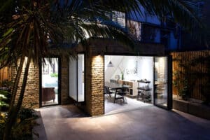 Modern home exterior with sleek aluminium bifold doors, revealing a stylish interior dining area. The space is surrounded by lush greenery and illuminated by warm lighting.