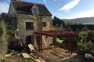 A charming stone house features a partially constructed red metal framework extending from the side, enhanced by sleek sliding aluminum doors. It's beautifully nestled within a lush garden and flanked by towering trees.