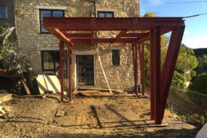 A steel frame structure is under construction in front of a stone house, set against a dirt ground and a clear blue sky, soon to be complemented by sleek sliding aluminium doors.