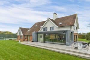 A large modern house with a sloped roof, patio, and lawn features slimline sliding doors. The patio offers outdoor seating and a view of a tennis court in the background.